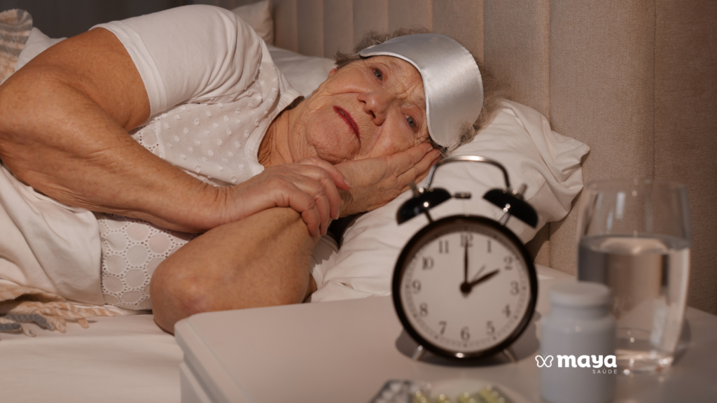 Idosa deitada na cama à noite, de olhos abertos e máscara de dormir na testa, ao lado de um despertador e remédios na mesa de cabeceira, ilustrando insônia.