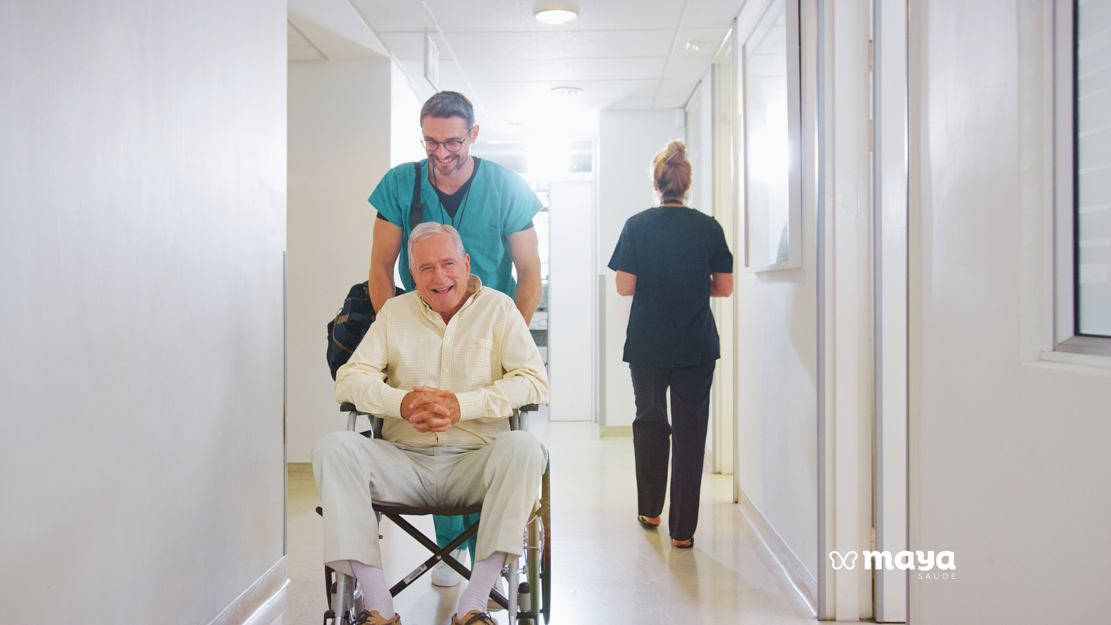 Enfermeiro sorridente conduzindo um homem idoso em cadeira de rodas pelo corredor do hospital, ilustrando a alta hospitalar do idoso.