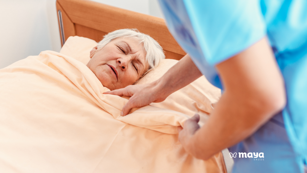 Idosa de cabelos brancos dormindo tranquila na cama enquanto uma cuidadora de uniforme azul a acompanha, ilustrando o cuidador noturno para idosos.