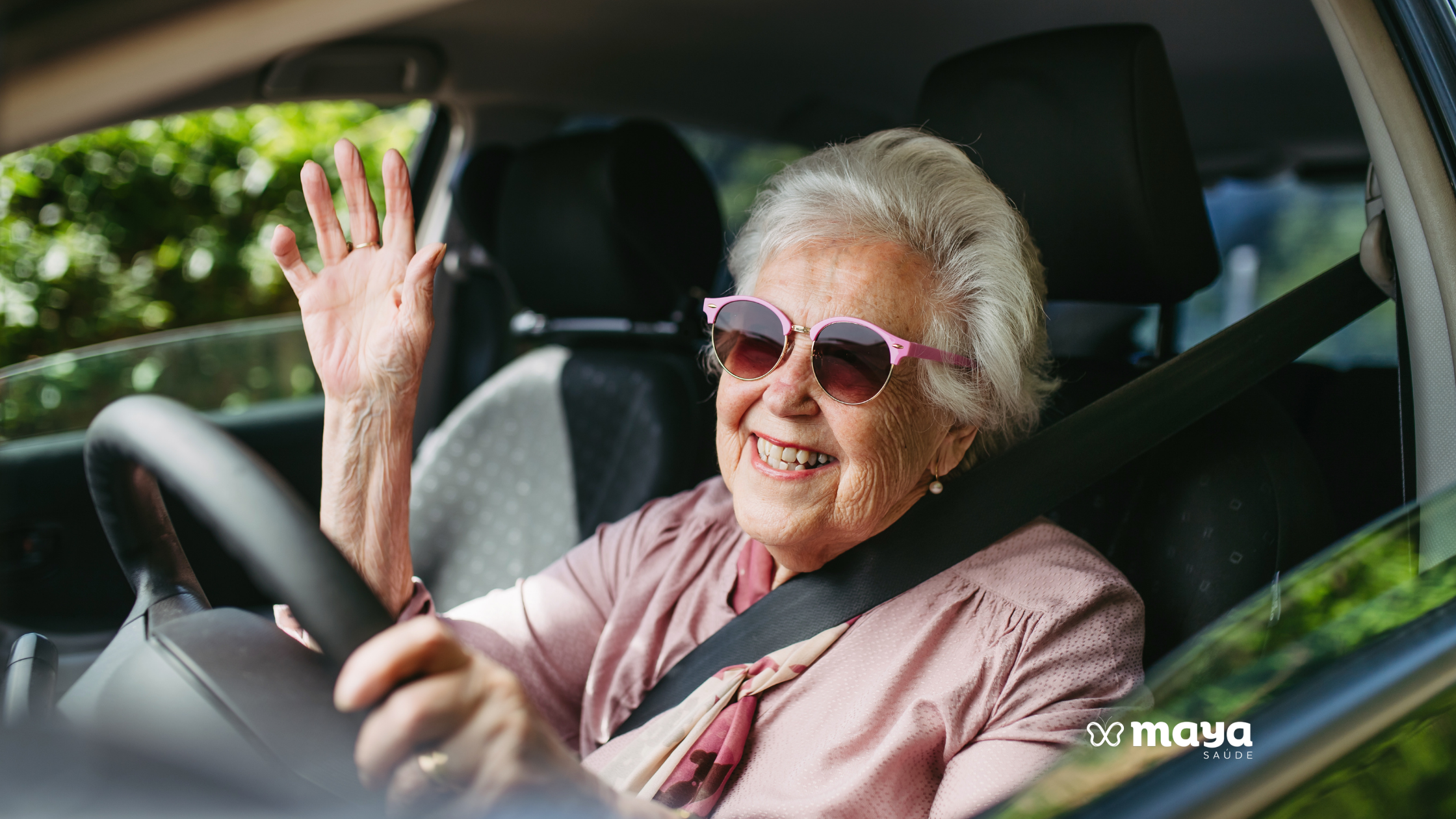 Idosa de cabelos brancos e óculos de sol rosa sorrindo ao volante do carro e acenando com a mão, ilustrando o tema dirigir na terceira idade.